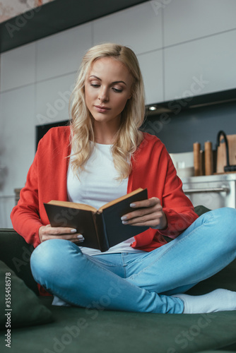 attentive young woman sitting on sofa with crossed legs and reading book