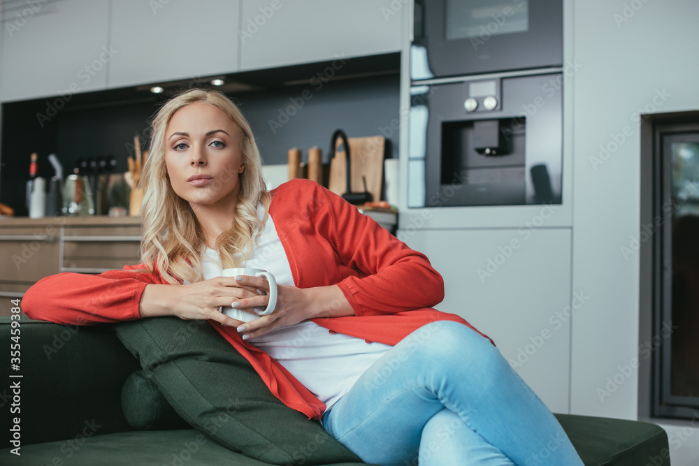 Obraz premium serious woman looking at camera while sitting on sofa with cup of tea