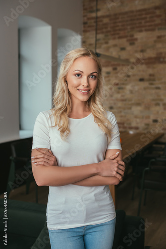 attractive blonde woman smiling at camera while standing at home with crossed arms
