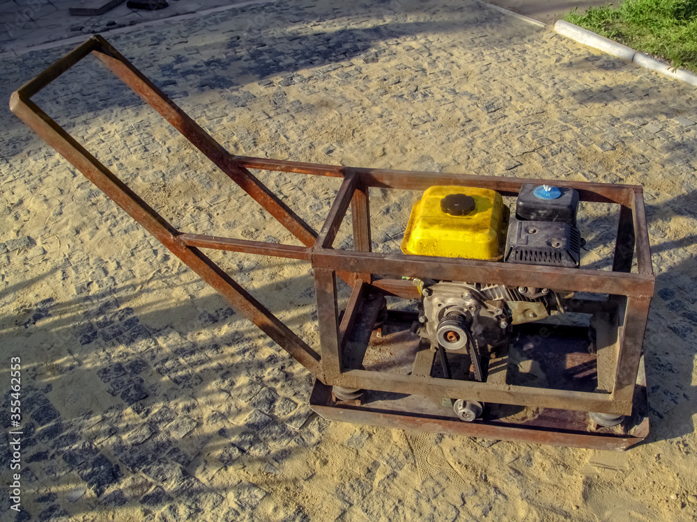 Homemade vibratory rammer device stands on the surface of a newly laid pavers on a sunny summer day. Concept of do-it-yourself vibro rammer with gasoline engine, self-laying paving slabs