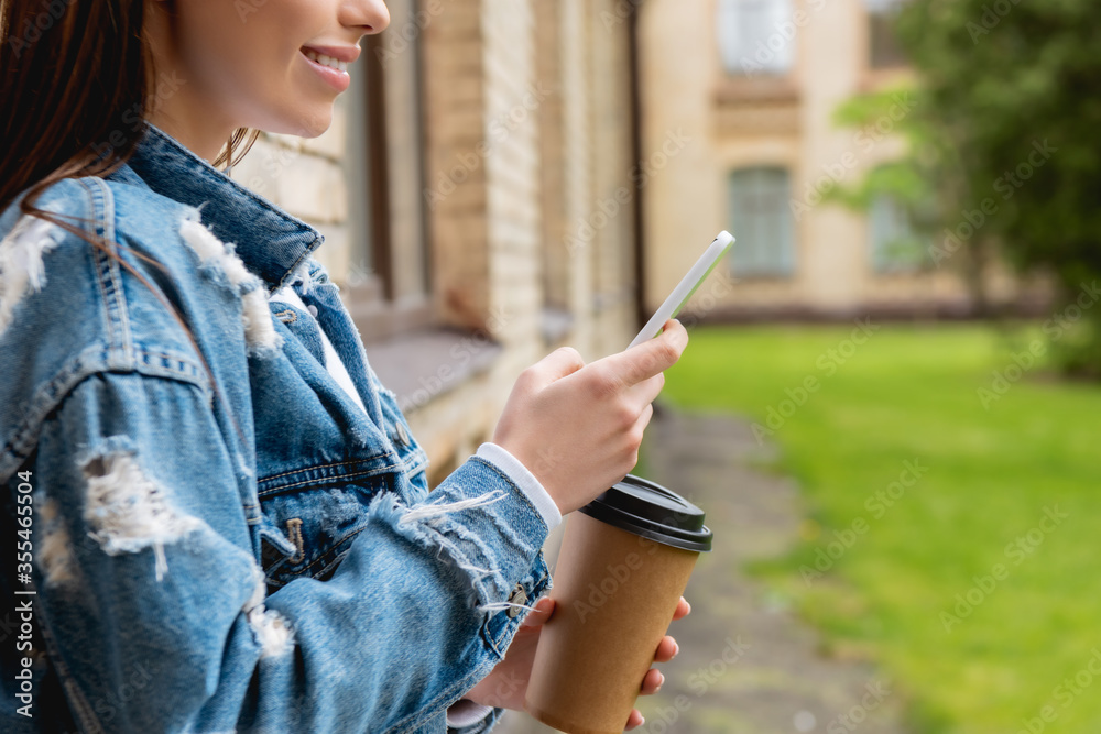 Cropped view of cheerful student using smartphone and holding paper cup near university campus Cropped view of cheerful student using smartphone and holding paper cup near university campus