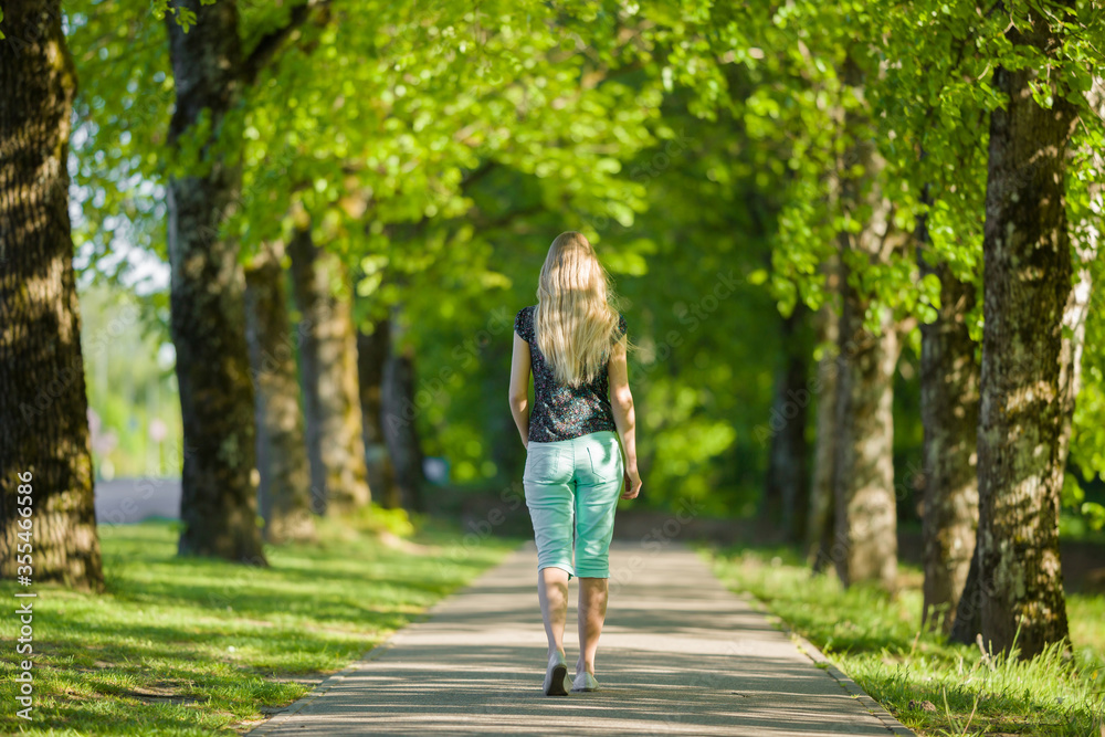 One young blonde woman slowly walking through green alley of trees in warm, sunny spring day. Shadow place. Spending time alone in nature. Peaceful atmosphere. Back view. One young blonde woman slowly walking through green alley of trees in warm, sunny spring day. Shadow place. Spending time alone in nature. Peaceful atmosphere. Back view.