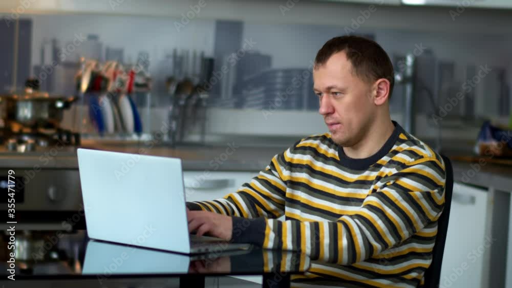 A man works at a laptop while sitting at home in the kitchen A man works at a laptop while sitting at home in the kitchen