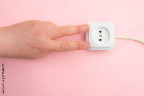 hand holds a metal plug near an electrical outlet. Rosette on a pink wall. Danger concept