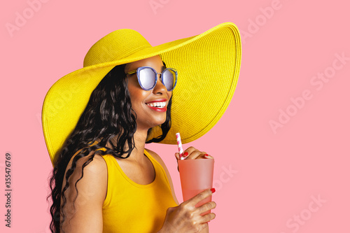 Profile portrait of a young smiling woman with yellow summer hat and sunglasses holding a drinking cup and paper straw