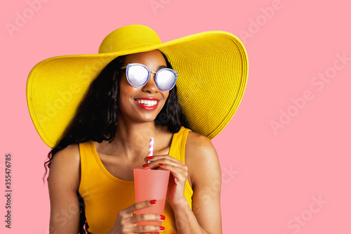 Portrait of a young smiling woman with yellow summer hat and sunglasses holding a drinking cup and paper straw