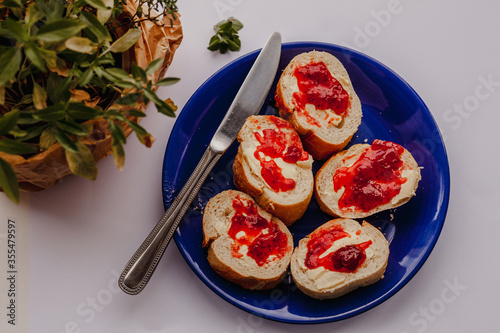 Slices of baguette bread with butter and strawberry jam on a blue plate with a metal butter knife on a pastel background