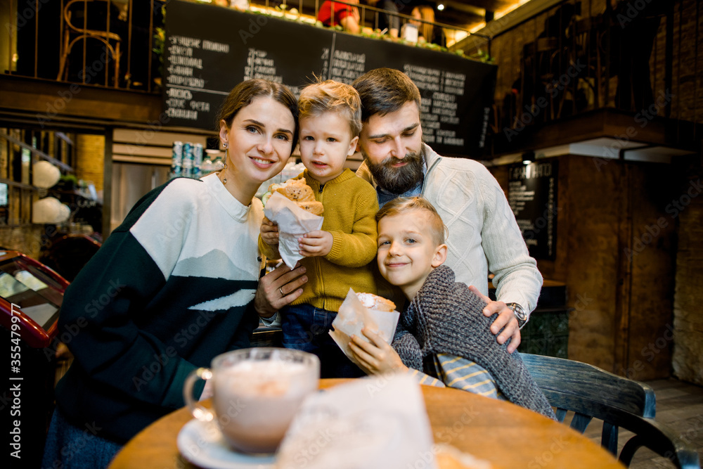 Joyful young smiling family with children sitting in cafe at the table, posing to camera with fresh tasty croissants in hands. Family breakfast in cafe