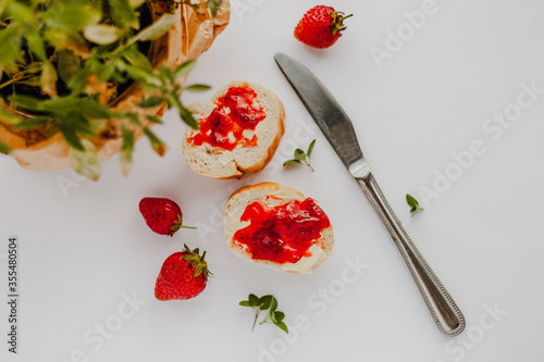 Slices of cut baguette bread with butter and strawberry jam, strawberries, metal butter knife and green thyme plant leaves on a pastel white background in a flat lay composition