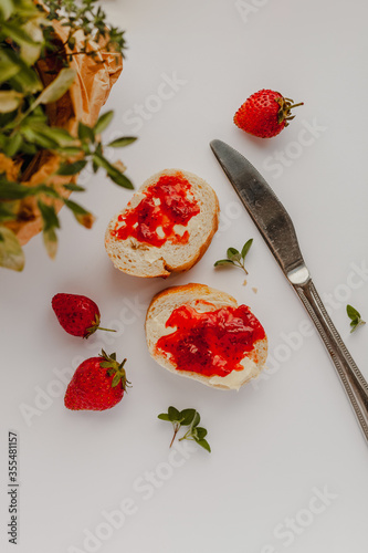 Slices of cut baguette bread with butter and strawberry jam, strawberries, metal butter knife and green thyme plant leaves on a pastel white background in a flat lay composition