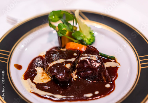 French main dish with stew of beef meat bourguignon simmered in red wine sauce with vegetables like carrots on a white and black dinner plate with golden rim.