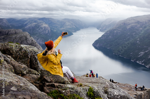 Happy blonde woman hiker in yellow raincoat sits on the top of the Preikestolen mountain (Preacher's Pulpit or Pulpit Rock) with a lot of tourists and shoots photos by phone on Lysefjord background