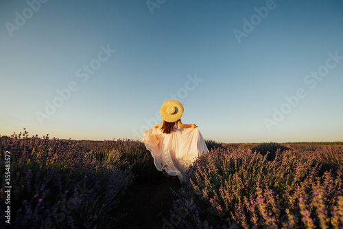 Back view of a pretty young girl in tender rose long dress with trendy straw hat running at the lavender field. Blue sky, sunny summer day. Copy space.