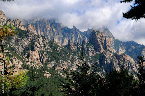 Pins Laricio et Aiguilles de bavella en Corse, sur fond de ciel bleu nuageux et nature sauvage et abrupte des rochers formant une montagne découpée au profil accidenté et dentelé.