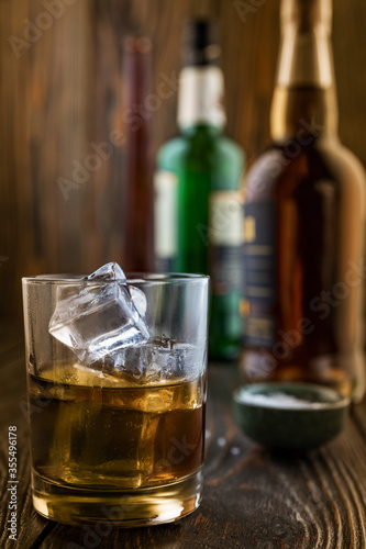 A glass of whiskey with ice, in the background are bottles on a wooden table of a bar counter, shallow depth of field, selective focus. The concept of alcoholic drinks in a roadside bar.