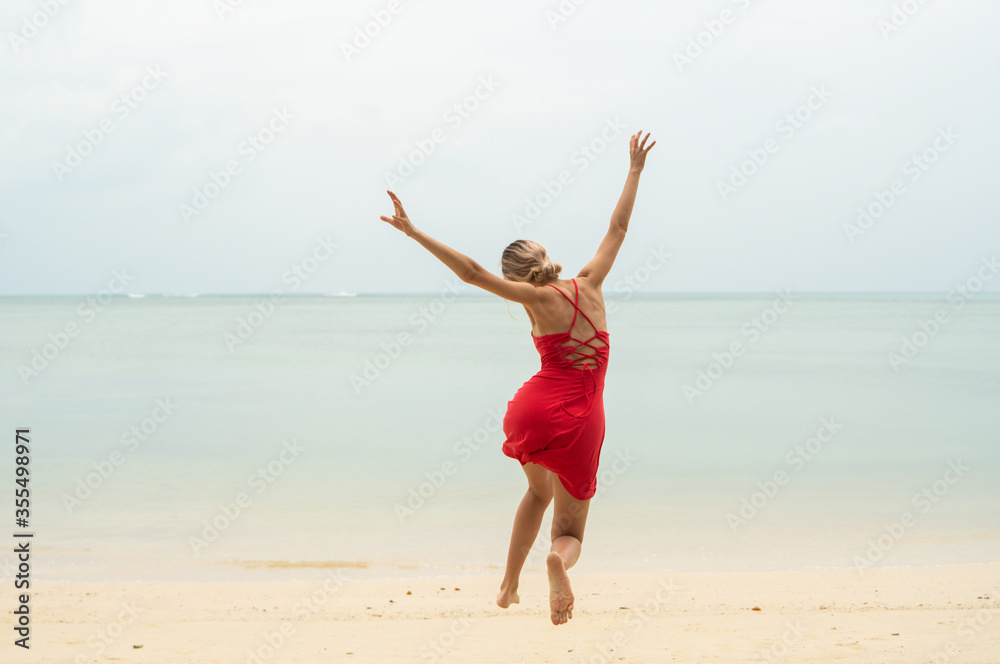 Young woman in a red dress runs and jumps on a white sand beach into tropical sea. Young happy woman running on the beach. Happiness to be on a sea or ocean. Vacation and travel concept.