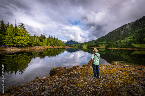 A Man standing at the shore in the morning enjoying the motionless water of the pacific ocean and its mirror like reflection in an inlet near Prince Rupert, British Columbia, Canada.
