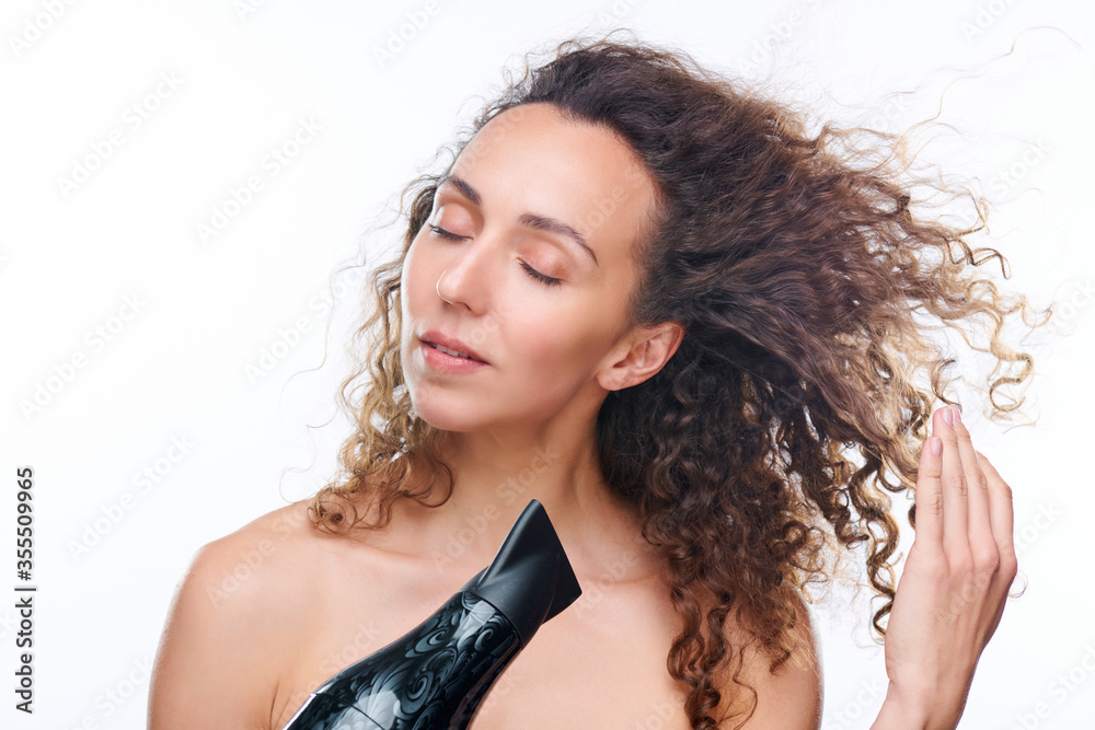 Pretty young woman with hair dryer drying her luxurious brown wavy clean hair