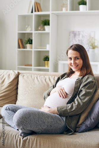 Happy young pregnant female in casualwear sitting among pillows on couch