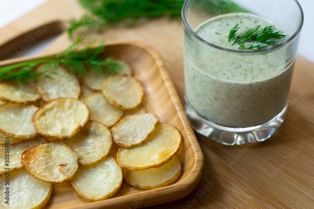 Dill and sour cream dip for homemade potato chips made vegan: healthy snack with dill, tahini, water, vinegar and lemon juice in a glass. Thinly sliced and baked potatoes in the back Dill and sour cream dip for homemade potato chips made vegan: healthy snack with dill, tahini, water, vinegar and lemon juice in a glass. Thinly sliced and baked potatoes in the back
