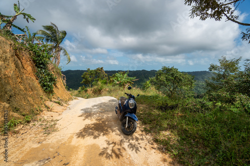 Blue scooter on the dirt road on a hill in tropical jungle with a scenic view on a mountains in a sunny day. Adventure and traveling concept.
