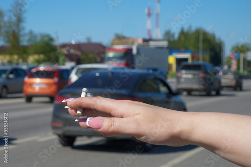  Hand of a girl with long nails and a cigarette on the background of a road with passing cars.