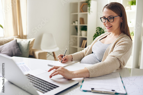 Happy young pregnant businesswoman looking at laptop display and making notes