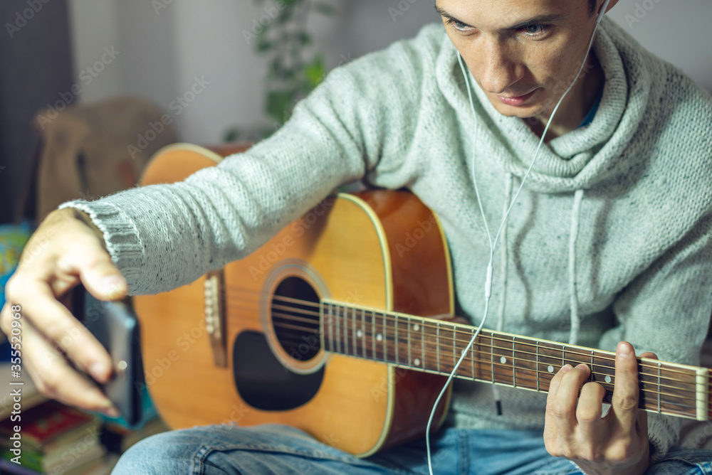 Obraz premium Young musician is learning to play acoustic guitar in an online lesson using phone app. Man is studying courses at home