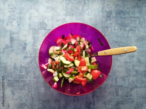 summer vegetable salad tomatoes cucumbers onions green radishes in a purple bowl on a background and a spoon