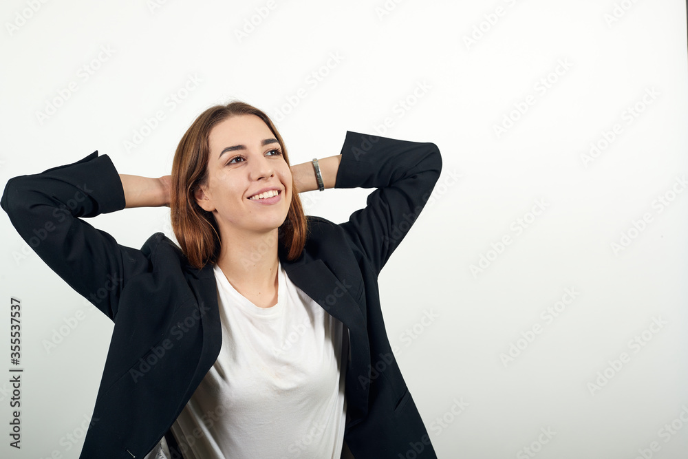 Taking time out to relax. Holding arms behind the head. Thinking about holidays, vacation. Enjoy lazy. Young attractive woman with brown hair in a light t-shirt and black jacket on white background Taking time out to relax. Holding arms behind the head. Thinking about holidays, vacation. Enjoy lazy. Young attractive woman with brown hair in a light t-shirt and black jacket on white background