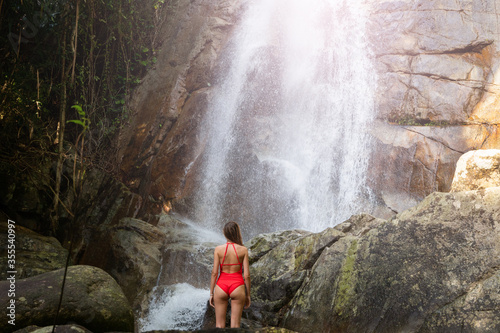 Beautiful young slim blonde woman with curly hair, wearing in red swimsuit is enjoy in lagoon of huge tropical waterfall in jungle. Travel concept. Sexy woman wearing red swimsuit in the jungle.