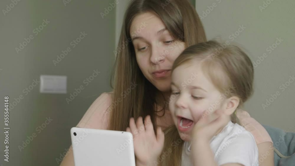 Mother with little cute daughter having fun and laughing using tablet. Funny games app. Mum with child take selfie, watch cartoons, make video call sitting on sofa at home. Mother with little cute daughter having fun and laughing using tablet. Funny games app. Mum with child take selfie, watch cartoons, make video call sitting on sofa at home.