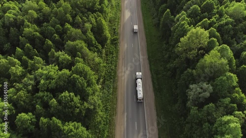 Gasoline tanker on asphalt road / White fuel semi truck fast driving on straight freeway in countryside dense forest at summer sunset / Aerial top down drone follow shot
