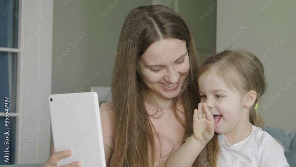 Mother with little cute daughter having fun and laughing using tablet. Funny games app. Mum with child take selfie, watch cartoons, make video call sitting on sofa at home. Mother with little cute daughter having fun and laughing using tablet. Funny games app. Mum with child take selfie, watch cartoons, make video call sitting on sofa at home.