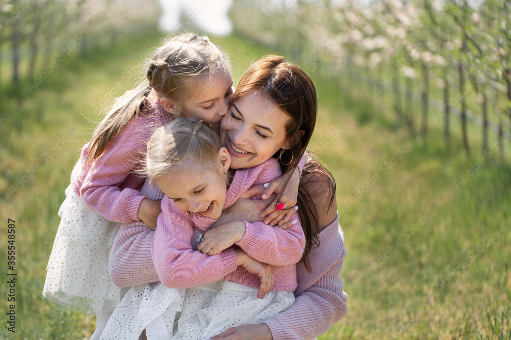 Portrait of a mother and her twin daughters in a blooming Apple orchard. Girls kiss their mother Portrait of a mother and her twin daughters in a blooming Apple orchard. Girls kiss their mother