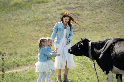 Young mother and her twin daughters feed a black cow in a meadow in the village