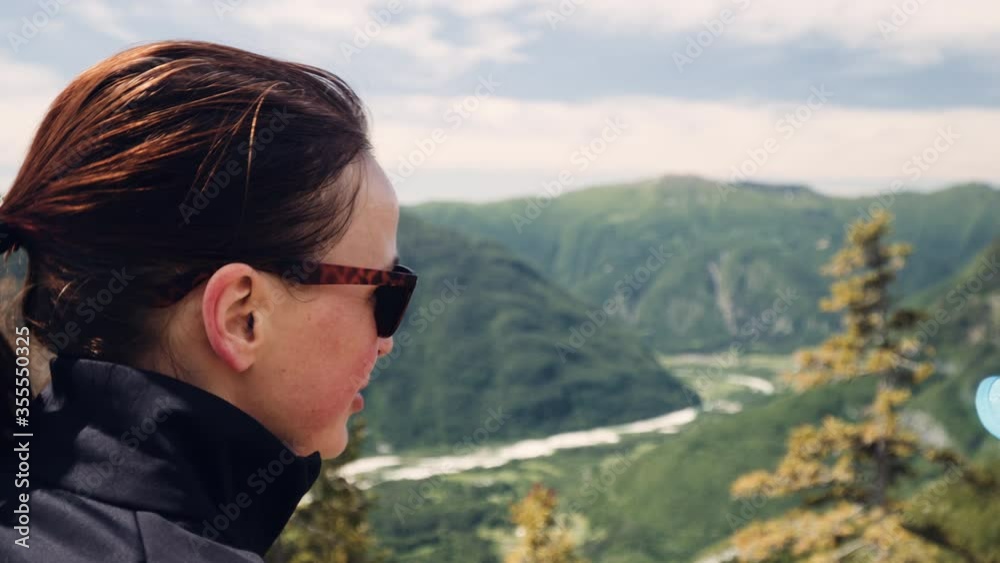Close up view, Caucasian woman with brown sunglasses talking to a friend of screen on top of the mountain, beautiful valley and river in background. Close up view, Caucasian woman with brown sunglasses talking to a friend of screen on top of the mountain, beautiful valley and river in background.