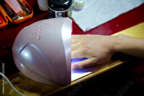 Closeup shoot . UV lamp for nail drying . Hand in UV lamp lights for nails on pink background. Minimal creative concept . Client with red varnish at manicure salon cairfully waiting when her nails