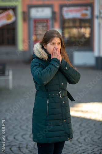 Young woman in warm winter coat coughing on the street