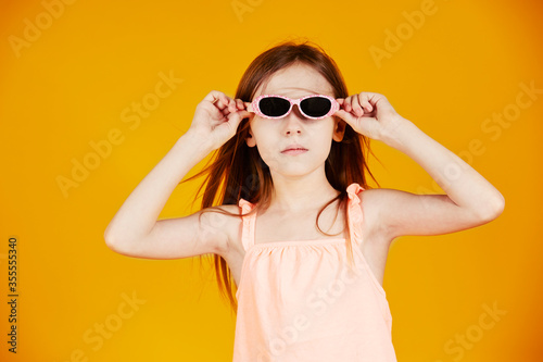 jolie jeune enfant fille caucasienne aux cheveux longs châtains portant des lunettes solaires en souriant sur le thème des vacances d'été sur fond jaune de studio