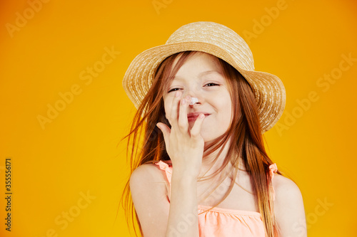 jolie jeune enfant fille caucasienne aux cheveux longs châtains mettant de la crème solaire sur son visage pour protéger sa peau sur fond jaune de studio