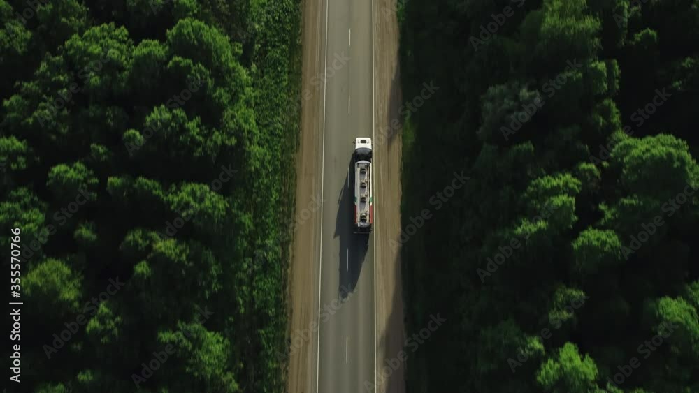 Gasoline tanker on asphalt road / White fuel semi truck fast driving on straight freeway in countryside dense forest at summer sunset / Aerial top down drone follow shot