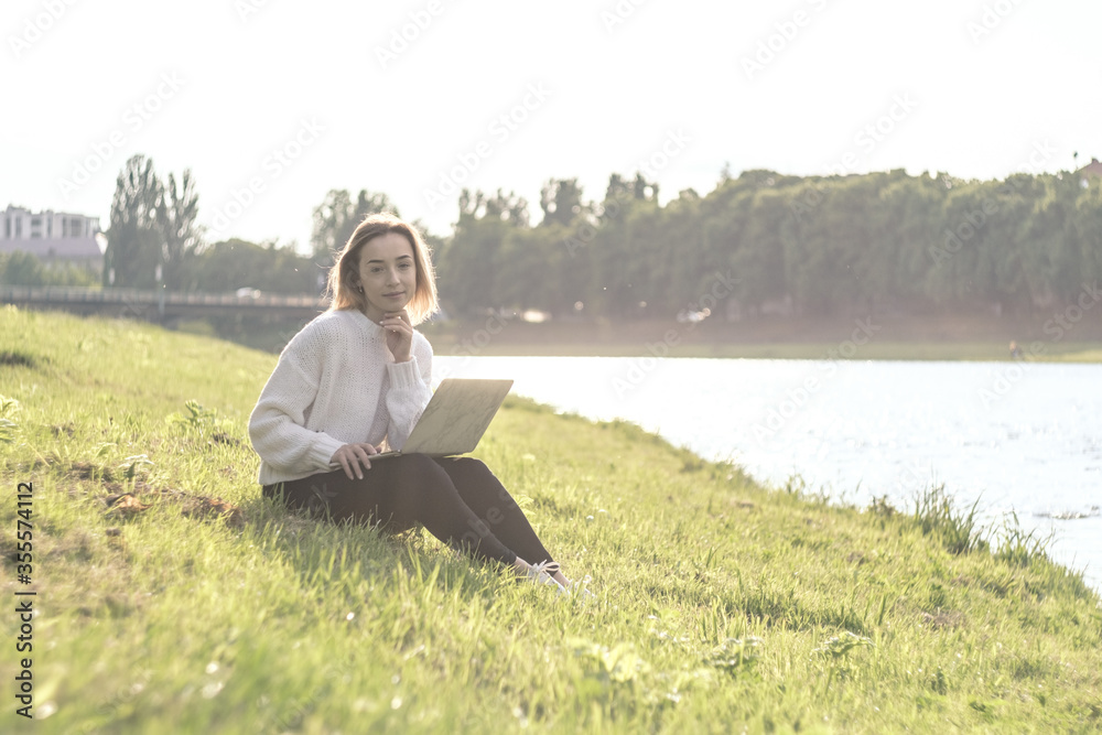 Fototapeta premium Blonde girl resting after working on a laptop on the grass by the river