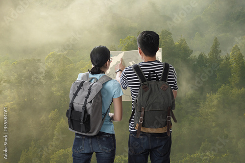 Travelers couple with the backpack on the top of mountain in the green forest and look searching direction on location map, Adventure and travel in the mountains region concept