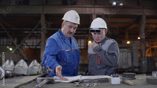 Two workers at heavy industry factory have conversation while looking at blueprint. Engineers explore and discuss project in heavy industry factory. Foundry for Metal Cast and Production.