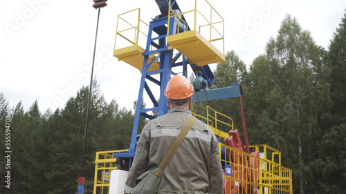 Oil field technical worker standing in front of crude oil pump unit. Engineer overseeing industrial oil pump jack. Oil industry concept.