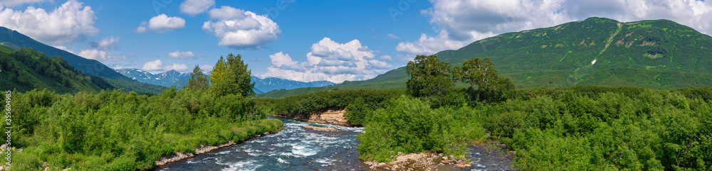 Panorama view of beautiful summer landscape - stream water of mountain river and green forest on hills along riverbank on sunny day with white clouds in blue sky. Headline summertime environment.
