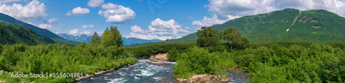 Panorama view of beautiful summer landscape - stream water of mountain river and green forest on hills along riverbank on sunny day with white clouds in blue sky. Headline summertime environment.