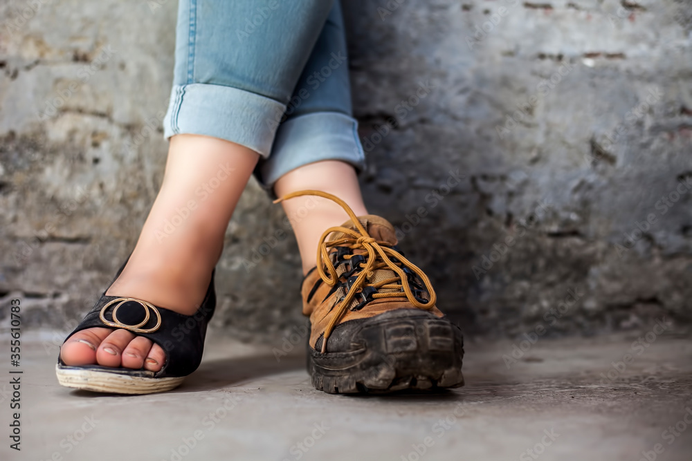 The elegant foot of young teenage girl wearing peep-toe sandals in one foot and hiking boots in another posing against a brick wall wearing blue denim jeans.