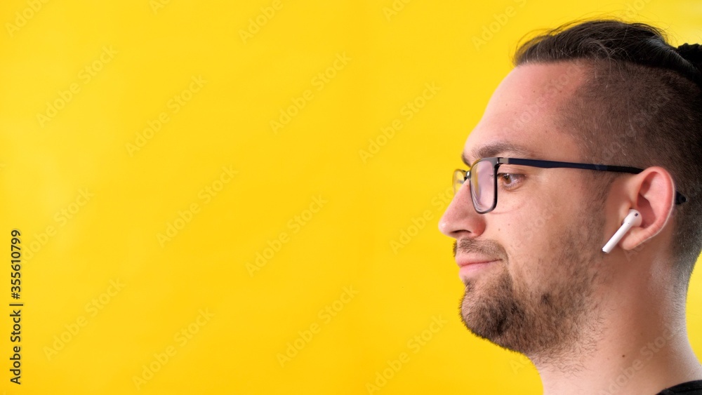 Obraz premium Displeased person man with glasses and headphones looks to the left side of the screen. Profile portrait of an attractive adult unshaven man on a yellow background. Upset emotional guy keeps calm